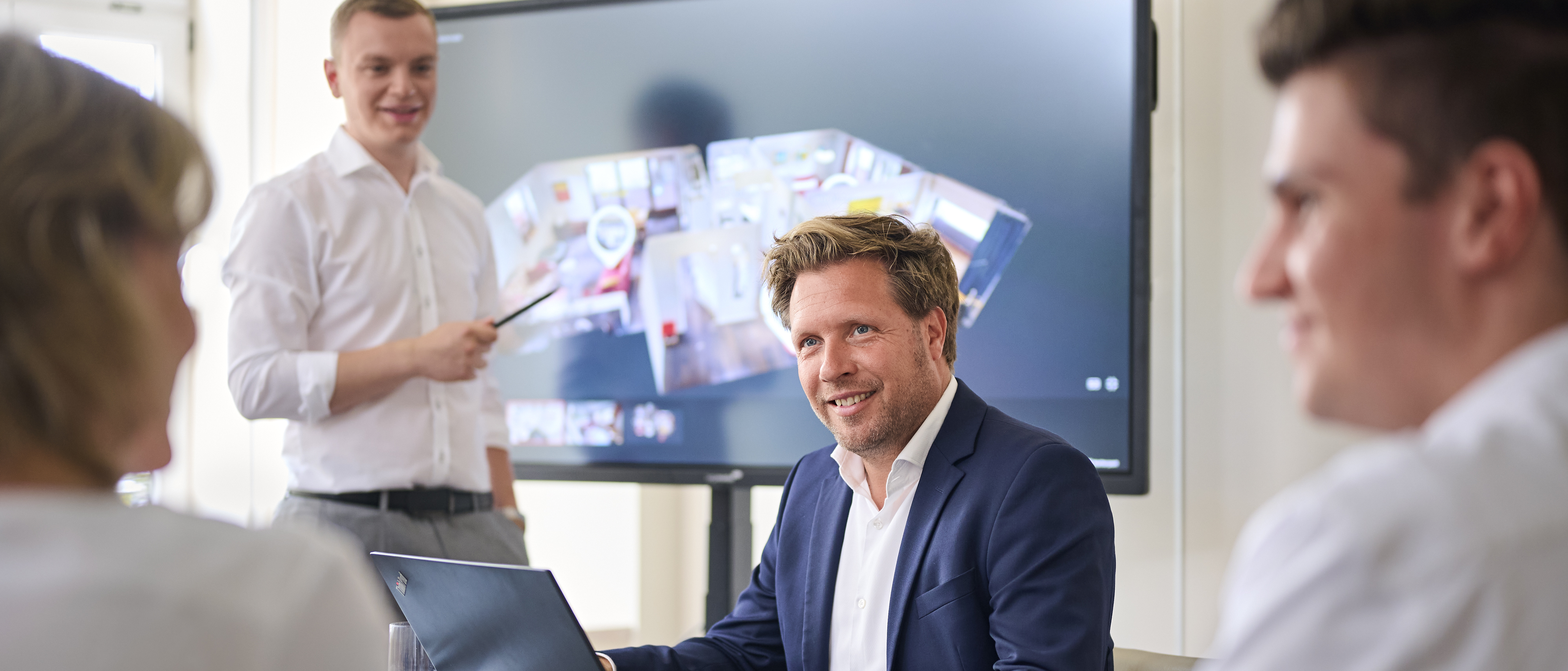 Drei Personen sitzen lachend an einem Tisch. Im Hintergrund steht eine männliche Person an einem TV-Bildschirm, auf dem ein Grundriss einer Immobilie abgebildet ist.
