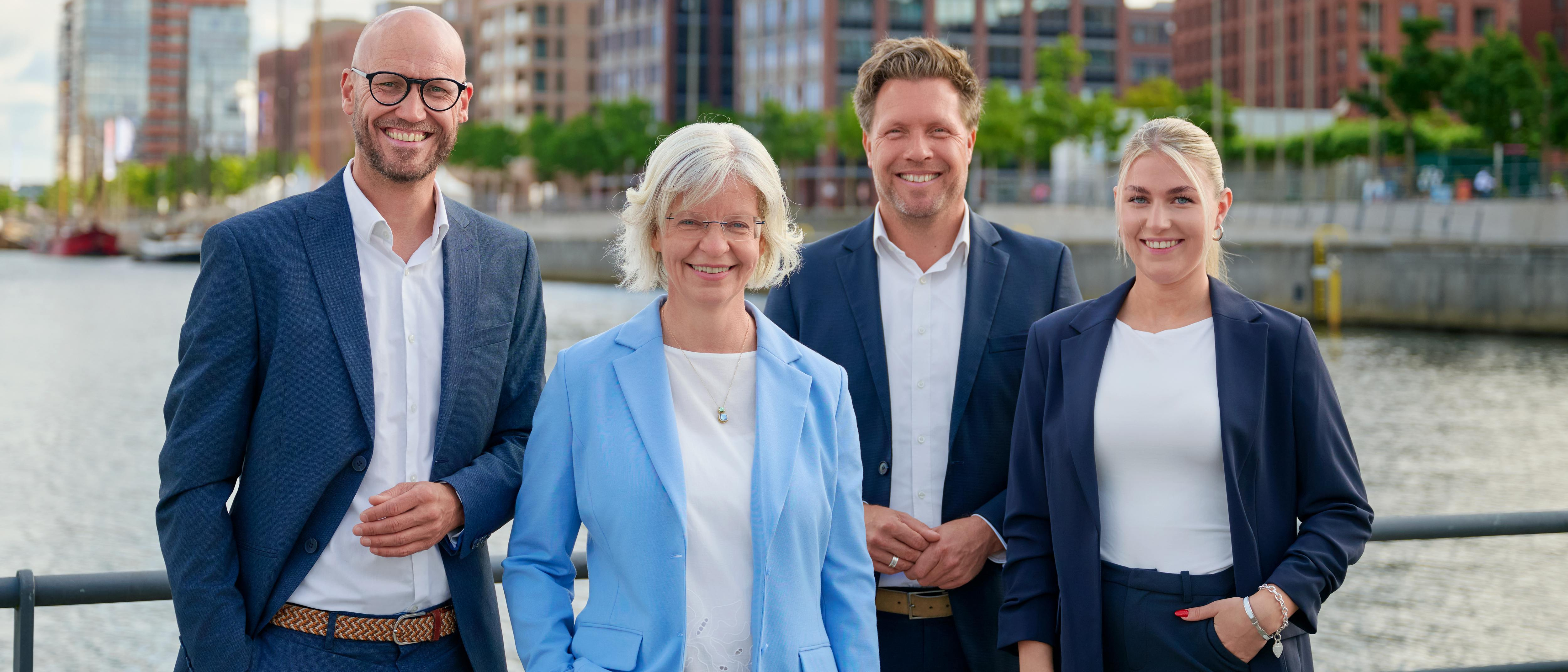 Vier Personen im Business-Outfit stehen am Wasser, an der Kieler Hörn, mit moderner Stadtarchitektur im Hintergrund. Von links nach rechts: Jan Wohlert, Wiebke Zauka, Jan Marin und Marisa Lebert.